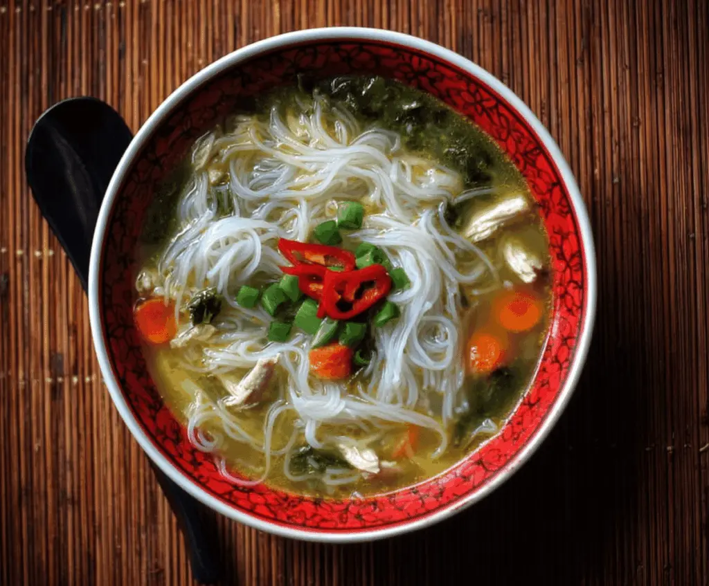 A steaming bowl of Chicken Rice Noodle Soup with tender chicken, rice noodles, fresh herbs, and sliced vegetables in a flavorful broth.