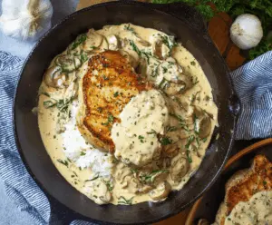 Juicy pork chops topped with rich, creamy garlic sauce served on a white plate, garnished with fresh herbs.